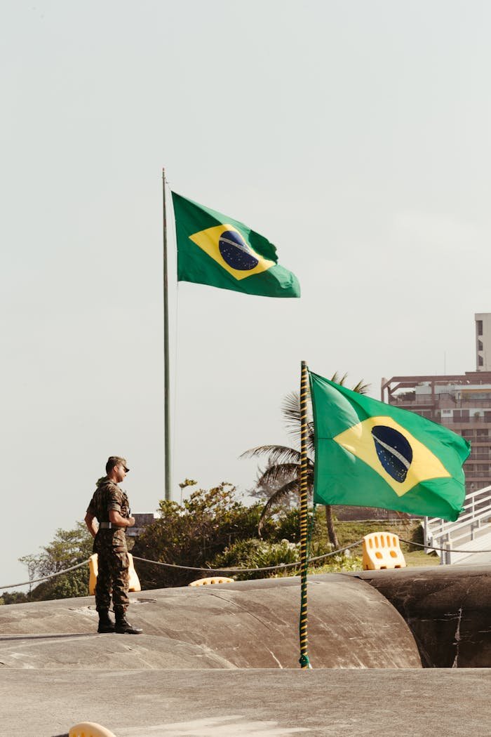 services-04 A soldier in uniform stands beside two Brazilian flags in an urban setting in Rio de Janeiro.